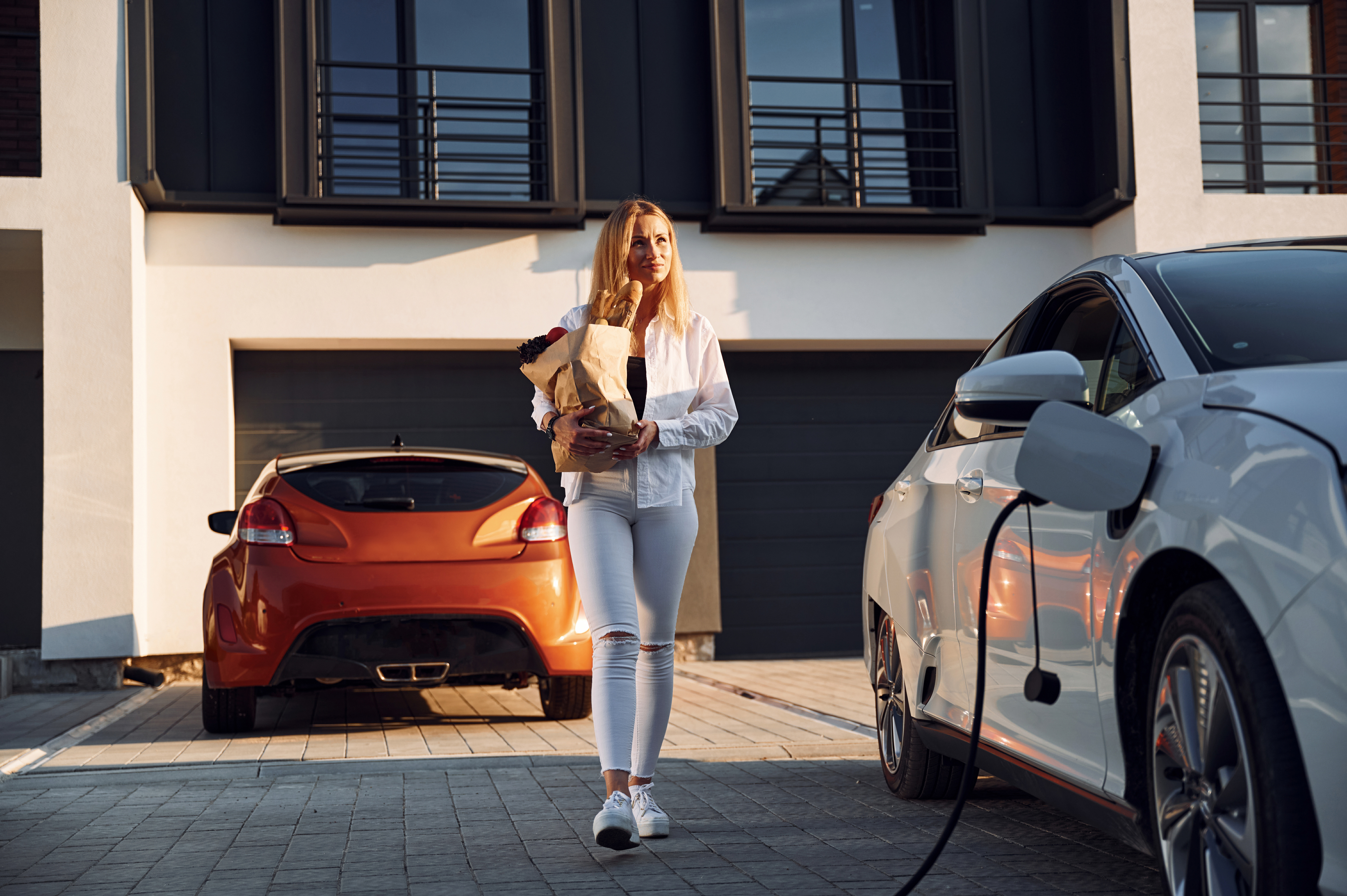 Holding shopping bag. Young woman in white clothes is with her electric car at daytime